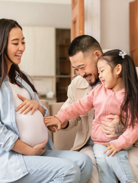 Second Pregnancy. Cheerful Asian Family Expecting Baby, Kid Daughter Touching Pregnant Mom's Belly Waiting For Little Brother Sitting On Sofa At Home. Expectation Happiness Concept